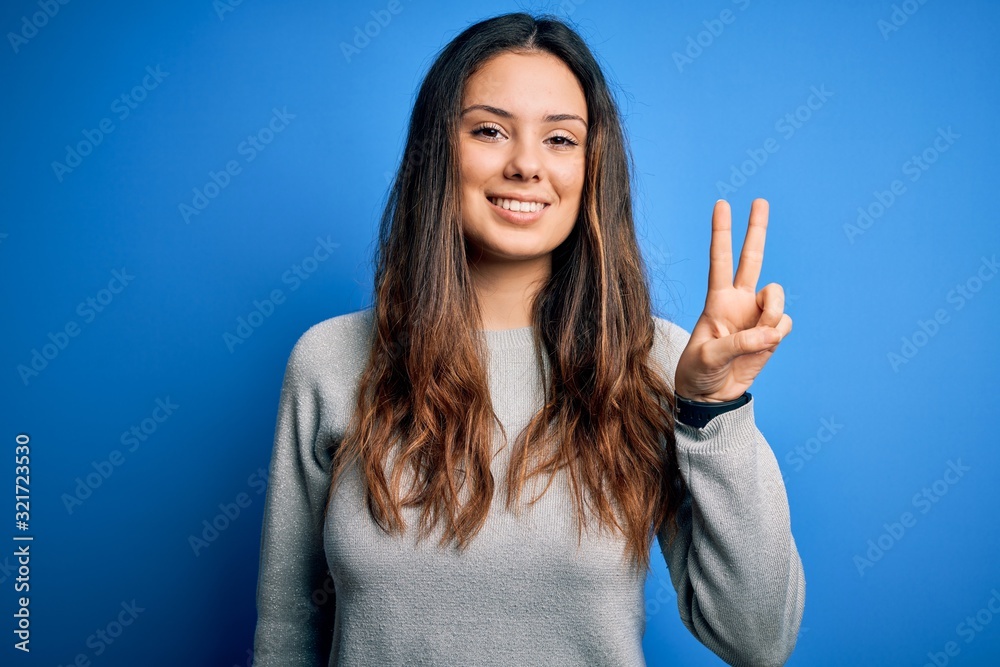 Young beautiful brunette woman wearing casual sweater standing over blue background smiling with happy face winking at the camera doing victory sign. Number two. Young beautiful brunette woman wearing casual sweater standing over blue background smiling with happy face winking at the camera doing victory sign. Number two.