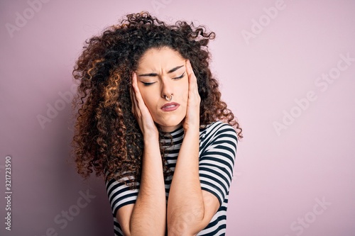 Young beautiful woman with curly hair and piercing wearing casual striped t-shirt with hand on head for pain in head because stress. Suffering migraine.