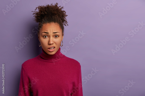 Human facial expressions concept. Displeased Afro woman smirks face and purses lips, reacts on hearing something unpleasant, wears casual jumper, models over purple wall with blank space aside photo
