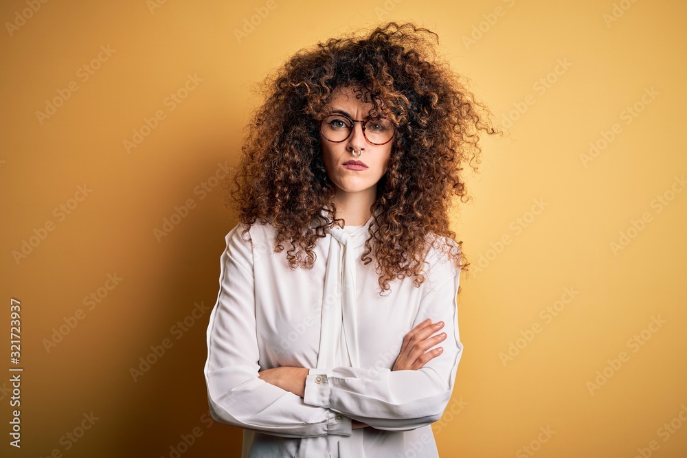Young beautiful brunette woman with curly hair and piercing wearing shirt and glasses skeptic and nervous, disapproving expression on face with crossed arms. Negative person. Young beautiful brunette woman with curly hair and piercing wearing shirt and glasses skeptic and nervous, disapproving expression on face with crossed arms. Negative person.