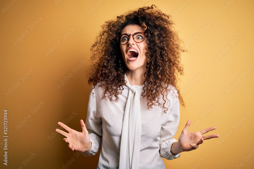 Young beautiful brunette woman with curly hair and piercing wearing shirt and glasses crazy and mad shouting and yelling with aggressive expression and arms raised. Frustration concept. Young beautiful brunette woman with curly hair and piercing wearing shirt and glasses crazy and mad shouting and yelling with aggressive expression and arms raised. Frustration concept.