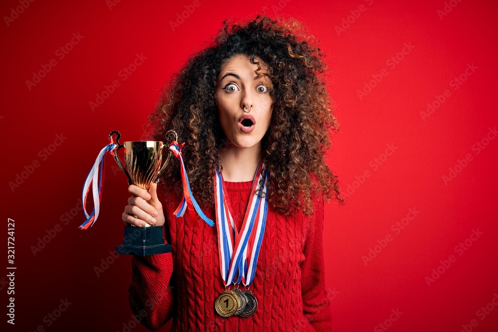 Young beautiful successful woman with curly hair and piercing holding trophy wearing medals scared in shock with a surprise face, afraid and excited with fear expression Young beautiful successful woman with curly hair and piercing holding trophy wearing medals scared in shock with a surprise face, afraid and excited with fear expression