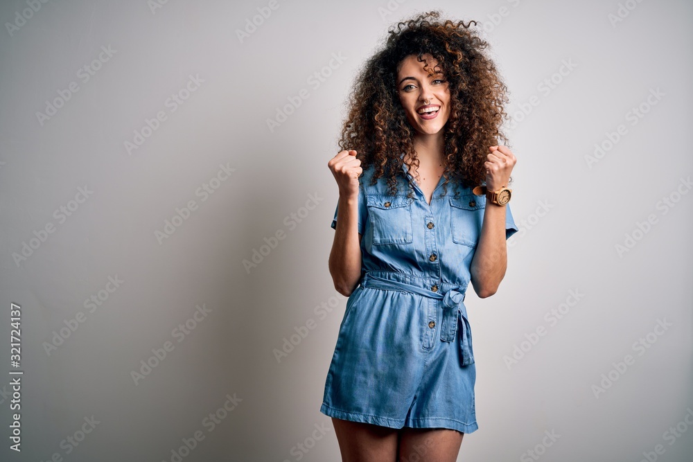 Young beautiful woman with curly hair and piercing wearing casual denim dress celebrating surprised and amazed for success with arms raised and open eyes. Winner concept.