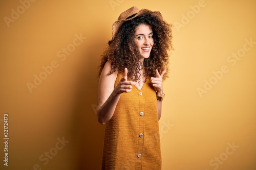 Beautiful brunette woman on vacation with curly hair and piercing wearing hat and dress pointing fingers to camera with happy and funny face. Good energy and vibes.