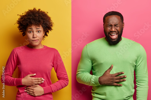 Fotografie, Obraz Horizontal shot of dark skinned couple touch stomachs, suffer from chronic gastritis, being hungry has displeased face expressions, isolated over yellow and pink background