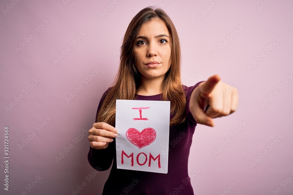 Young beautiful woman holding paper with love mom message celebrating mothers day pointing with finger to the camera and to you, hand sign, positive and confident gesture from the front Young beautiful woman holding paper with love mom message celebrating mothers day pointing with finger to the camera and to you, hand sign, positive and confident gesture from the front