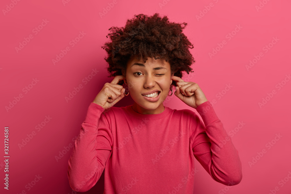 Portrait of dark skinned model plugs ears and ignores loud noise, clenches teeth, winks eyes, avoids unpleasant sound on party, wears casual rosy jumper, isolated over vibrant pink background Portrait of dark skinned model plugs ears and ignores loud noise, clenches teeth, winks eyes, avoids unpleasant sound on party, wears casual rosy jumper, isolated over vibrant pink background