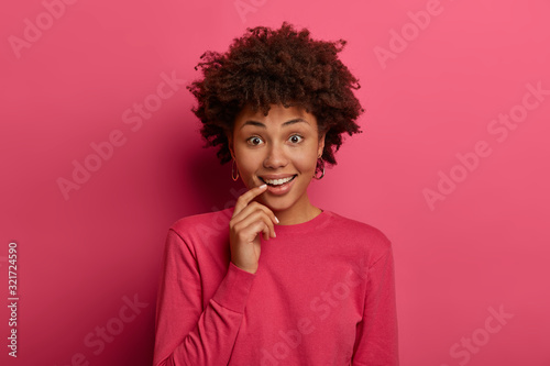 Curious dark skinned woman has glad expression  keeps index finger near lips  hears interesting suggestion or information  wears crimson jumper  isolated on pink wall. Human facial expressions concept