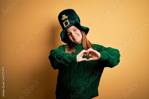 Young beautiful brunette woman wearing green hat on st patricks day celebration smiling in love doing heart symbol shape with hands. Romantic concept.