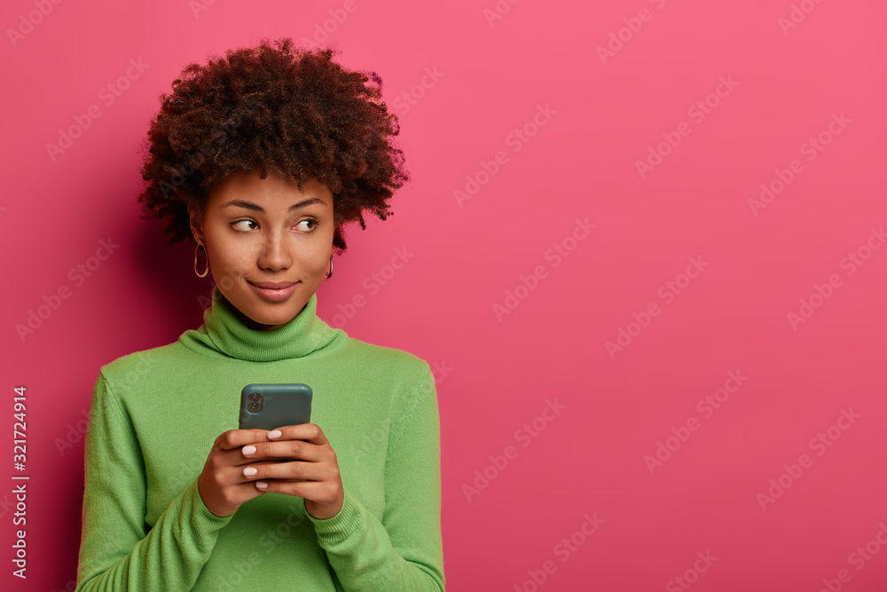 Thoughtful curly hipster girl creats post on own web page, uses mobile phone, being always in touch, reads comments, concentrated aside, copy space on vibrant pink background, reads website blog Thoughtful curly hipster girl creats post on own web page, uses mobile phone, being always in touch, reads comments, concentrated aside, copy space on vibrant pink background, reads website blog
