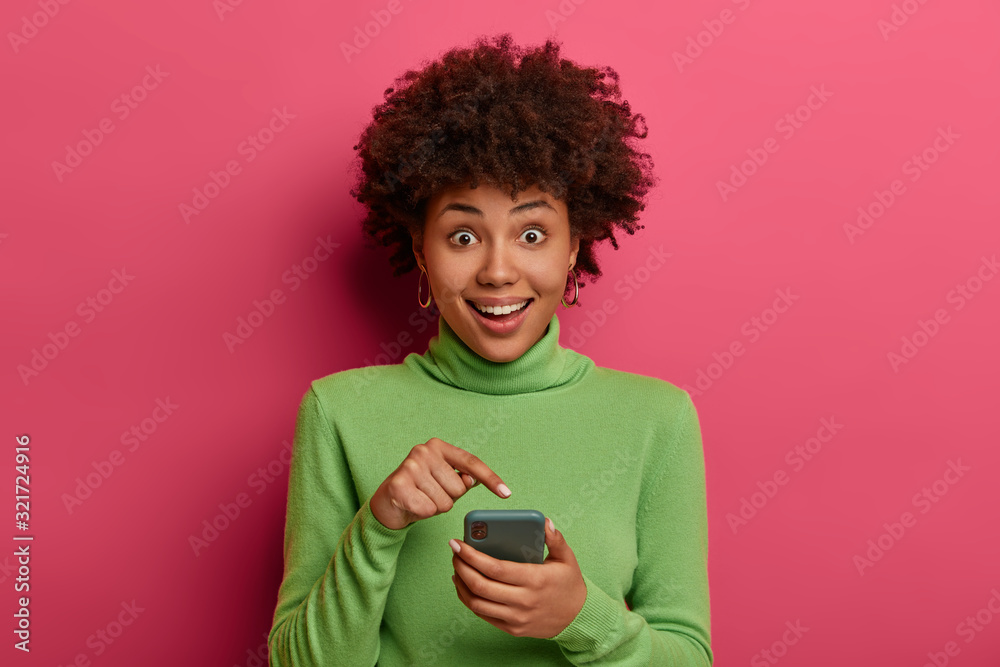 Excited happy ethnic female points at smartphone display, glad to receive romantic date invitation, wears casual green poloneck, sends new just made selfie on web page, stands in vivid studio Excited happy ethnic female points at smartphone display, glad to receive romantic date invitation, wears casual green poloneck, sends new just made selfie on web page, stands in vivid studio