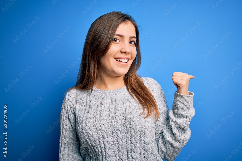 Beautiful young woman wearing casual wool sweater standing over blue isolated background smiling with happy face looking and pointing to the side with thumb up.