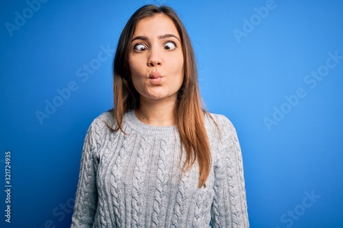 Beautiful young woman wearing casual wool sweater standing over blue isolated background making fish face with lips, crazy and comical gesture. Funny expression. © Krakenimages.com