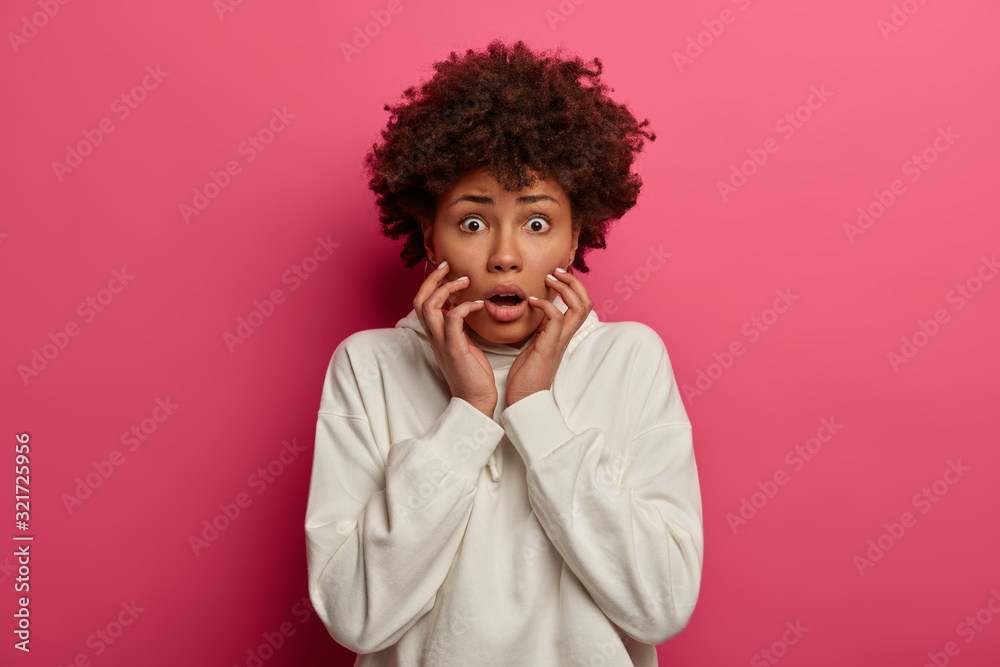 Nervous woman gasps from fear, keeps hands near opened mouth, afraids of something terrible, has worried expression, expresses negative emotions, wears white sweatshirt. People, reaction concept Nervous woman gasps from fear, keeps hands near opened mouth, afraids of something terrible, has worried expression, expresses negative emotions, wears white sweatshirt. People, reaction concept