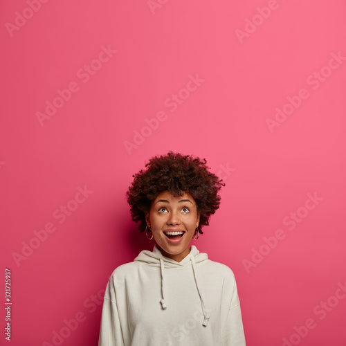 Happy ethnic girl concentrated above on ceiling, sees amazing thing upwards, smiles joyfully, has funny curious look up, wears casual clothes, isolated over rosy wall with copy space. Wow, interesting