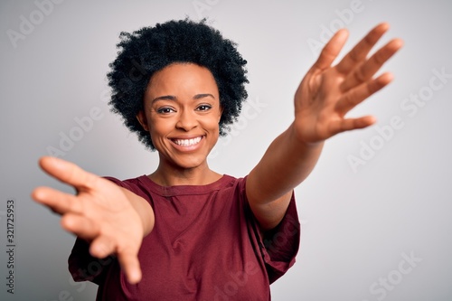 Young beautiful African American afro woman with curly hair wearing casual t-shirt standing looking at the camera smiling with open arms for hug. Cheerful expression embracing happiness.