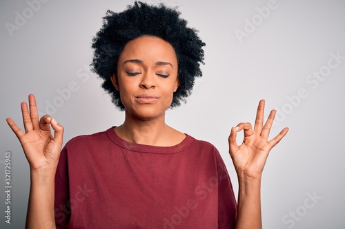 Young beautiful African American afro woman with curly hair wearing casual t-shirt standing relax and smiling with eyes closed doing meditation gesture with fingers. Yoga concept.