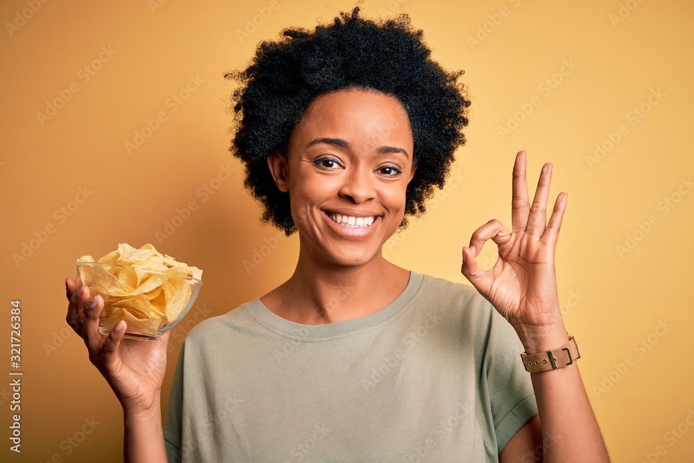 Young African American afro woman with curly hair holding bowl with chips potatoes doing ok sign with fingers, excellent symbol Young African American afro woman with curly hair holding bowl with chips potatoes doing ok sign with fingers, excellent symbol