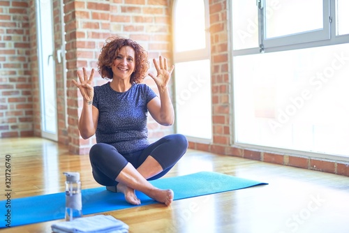 Middle age beautiful sportswoman wearing sportswear sitting on mat practicing yoga at home showing and pointing up with fingers number nine while smiling confident and happy.
