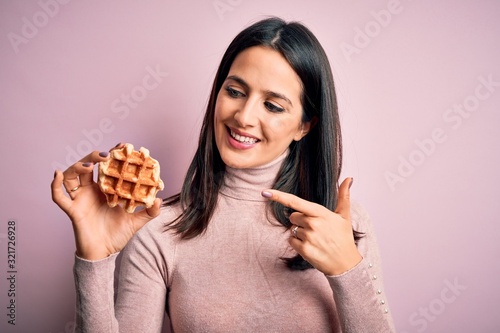 Young brunette woman with blue eyes eating sweet waffle for breakfast over white background very happy pointing with hand and finger