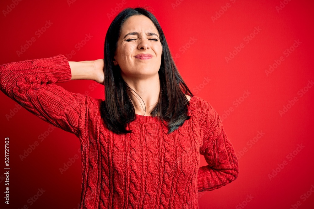 Young brunette woman with blue eyes wearing casual sweater over isolated red background Suffering of neck ache injury, touching neck with hand, muscular pain Young brunette woman with blue eyes wearing casual sweater over isolated red background Suffering of neck ache injury, touching neck with hand, muscular pain