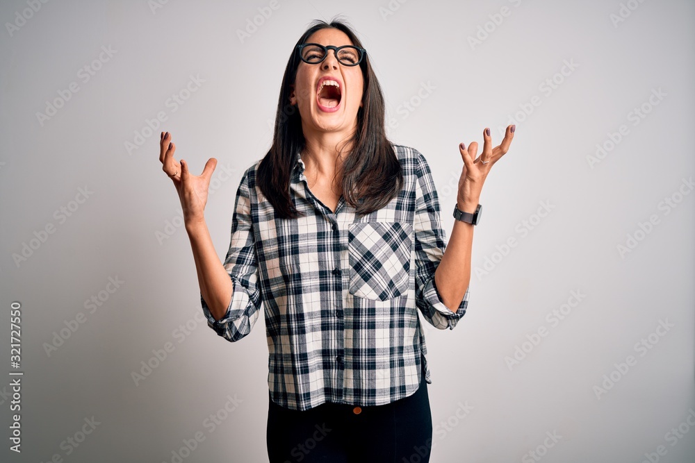 Young brunette woman with blue eyes wearing casual shirt and glasses over white background crazy and mad shouting and yelling with aggressive expression and arms raised. Frustration concept. Young brunette woman with blue eyes wearing casual shirt and glasses over white background crazy and mad shouting and yelling with aggressive expression and arms raised. Frustration concept.