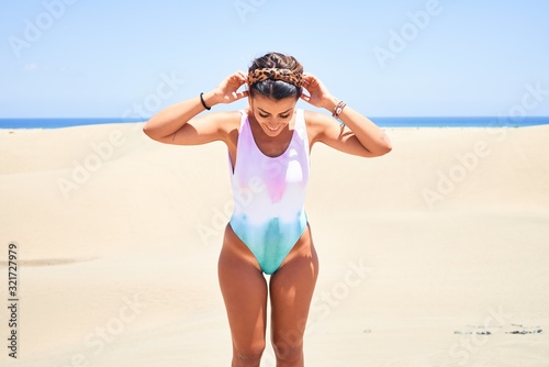 Young beautiful girl on vacation wearing swimsuit smiling happy and confident. Standing with a smile on face looking down wirh hands on head at beach