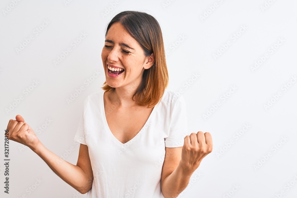Beautiful redhead woman wearing casual white t-shirt over isolated background very happy and excited doing winner gesture with arms raised, smiling and screaming for success. Celebration concept.