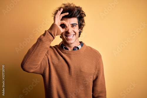 Young handsome man wearing casual shirt and sweater over isolated yellow background doing ok gesture with hand smiling, eye looking through fingers with happy face. © Krakenimages.com