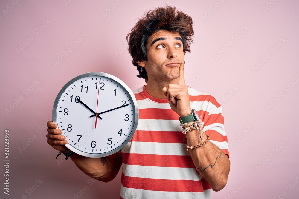 Young handsome man holding big clock standing over isolated pink background serious face thinking about question, very confused idea Young handsome man holding big clock standing over isolated pink background serious face thinking about question, very confused idea