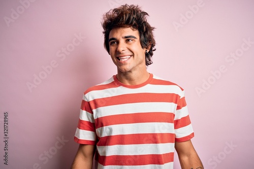 Young handsome man wearing striped casual t-shirt standing over isolated pink background looking away to side with smile on face, natural expression. Laughing confident.