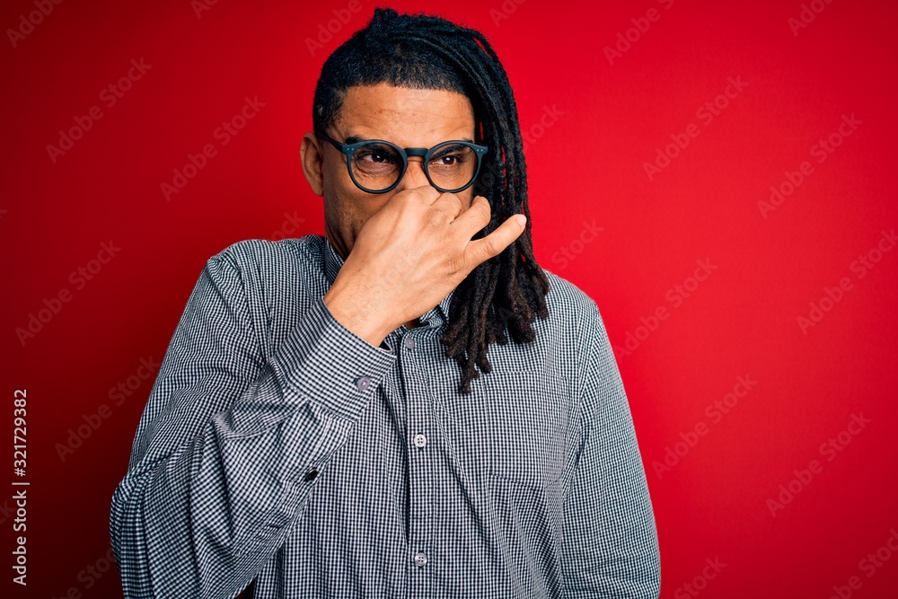 Fototapeta premium Young handsome african american man with dreadlocks wearing casual shirt and glasses smelling something stinky and disgusting, intolerable smell, holding breath with fingers on nose. Bad smell