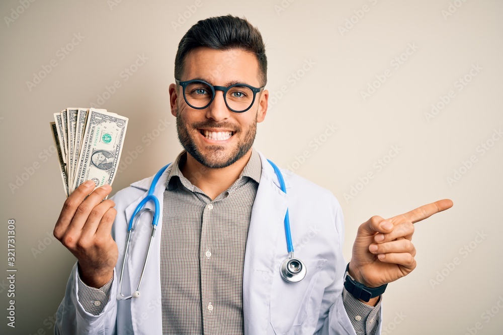 Young doctor man holding a bunch of one dollar banknotes over isolated background very happy pointing with hand and finger to the side Young doctor man holding a bunch of one dollar banknotes over isolated background very happy pointing with hand and finger to the side