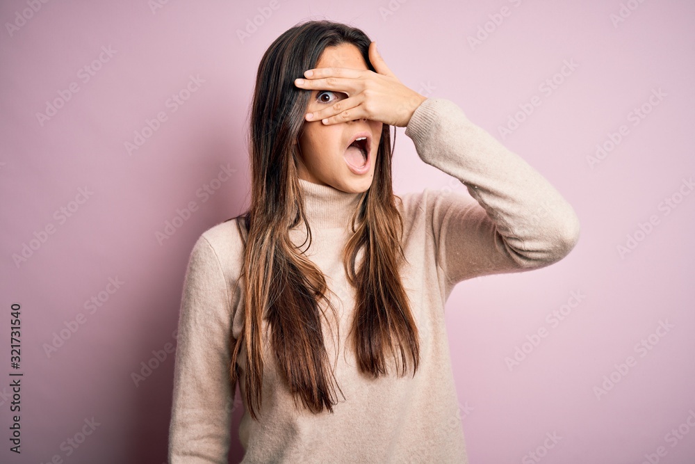 Young beautiful girl wearing casual turtleneck sweater standing over isolated pink background peeking in shock covering face and eyes with hand, looking through fingers with embarrassed expression. Young beautiful girl wearing casual turtleneck sweater standing over isolated pink background peeking in shock covering face and eyes with hand, looking through fingers with embarrassed expression.