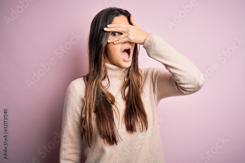 Young beautiful girl wearing casual turtleneck sweater standing over isolated pink background peeking in shock covering face and eyes with hand, looking through fingers with embarrassed expression.