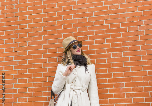 Portrait of an attractive and pretty blonde woman with hat, black glasses and white coat in front of a pair of bricks