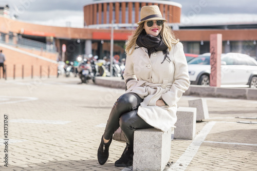 Very attractive blonde caucasian woman with hat and white coat smiles sitting by a large station