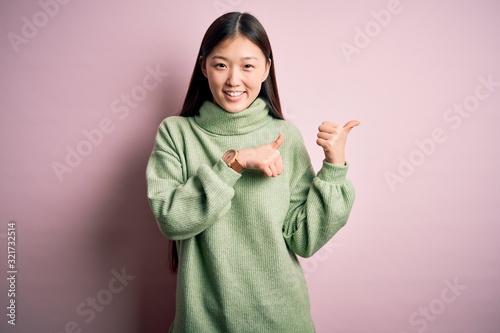 Young beautiful asian woman wearing green winter sweater over pink solated background Pointing to the back behind with hand and thumbs up, smiling confident