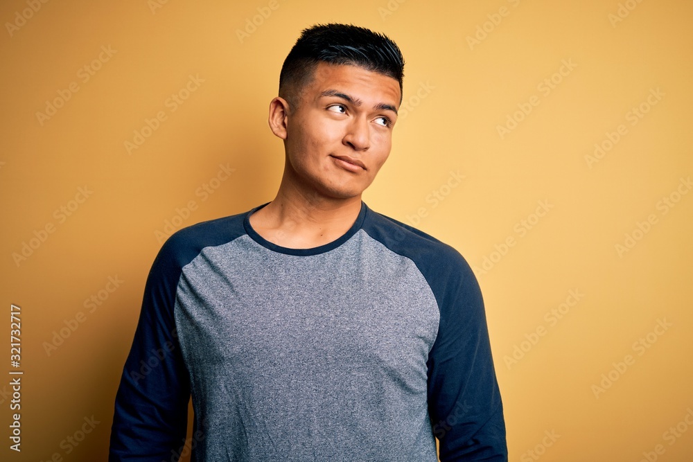 Young handsome latin man wearing casual t-shirt standing over yellow background smiling looking to the side and staring away thinking. Young handsome latin man wearing casual t-shirt standing over yellow background smiling looking to the side and staring away thinking.