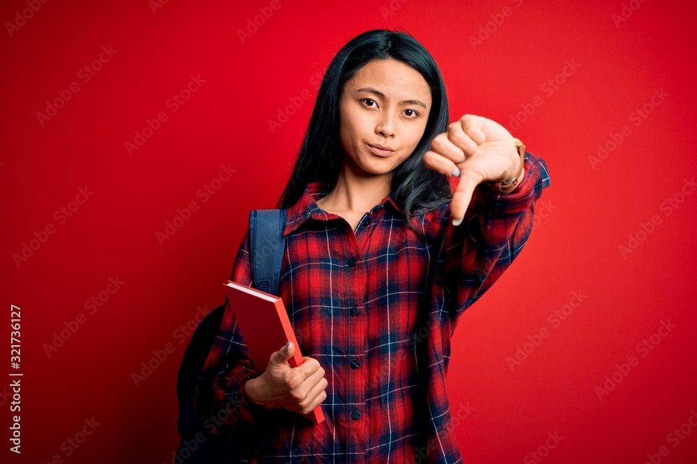 Young beautiful chinese student woman holding book standing over isolated pink background with angry face, negative sign showing dislike with thumbs down, rejection concept Young beautiful chinese student woman holding book standing over isolated pink background with angry face, negative sign showing dislike with thumbs down, rejection concept