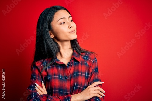 Young beautiful chinese woman wearing casual shirt over isolated red background looking to the side with arms crossed convinced and confident