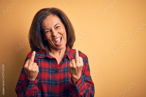 Middle age beautiful woman wearing casual shirt standing over isolated yellow background Showing middle finger doing fuck you bad expression, provocation and rude attitude. Screaming excited