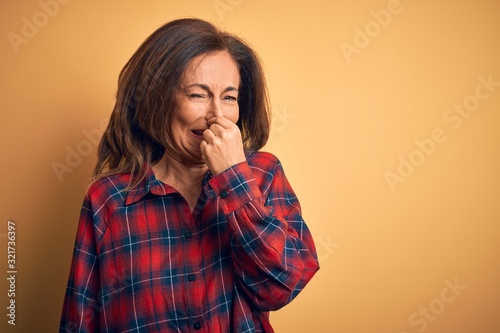 Middle age beautiful woman wearing casual shirt standing over isolated yellow background smelling something stinky and disgusting, intolerable smell, holding breath with fingers on nose. Bad smell