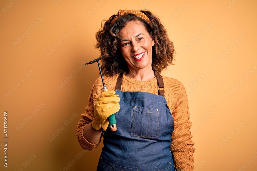 Middle age gardener woman wearing apron holding rake over isolated yellow background with a happy face standing and smiling with a confident smile showing teeth Middle age gardener woman wearing apron holding rake over isolated yellow background with a happy face standing and smiling with a confident smile showing teeth
