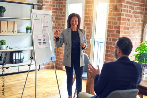 Two middle age business workers smiling happy and confidentt working together with smile on face. Woman standing doing work presentation using board and charts at the office photo