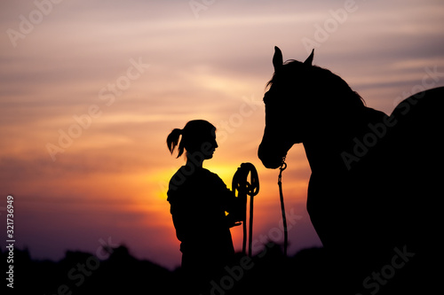 The girl near to a horse standing in front of a beautiful sunset. Silhouette of a woman and a horse. Human holding the horse by the rope.