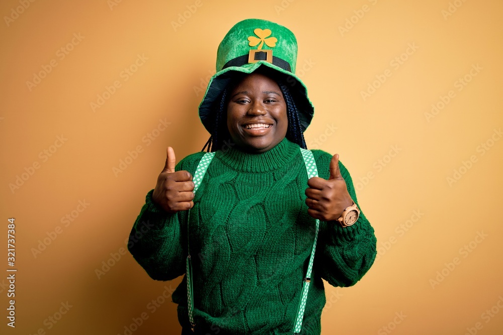 Plus size african american woman with braids wearing green hat with clover on st patricks day success sign doing positive gesture with hand, thumbs up smiling and happy. Cheerful expression and winner Plus size african american woman with braids wearing green hat with clover on st patricks day success sign doing positive gesture with hand, thumbs up smiling and happy. Cheerful expression and winner
