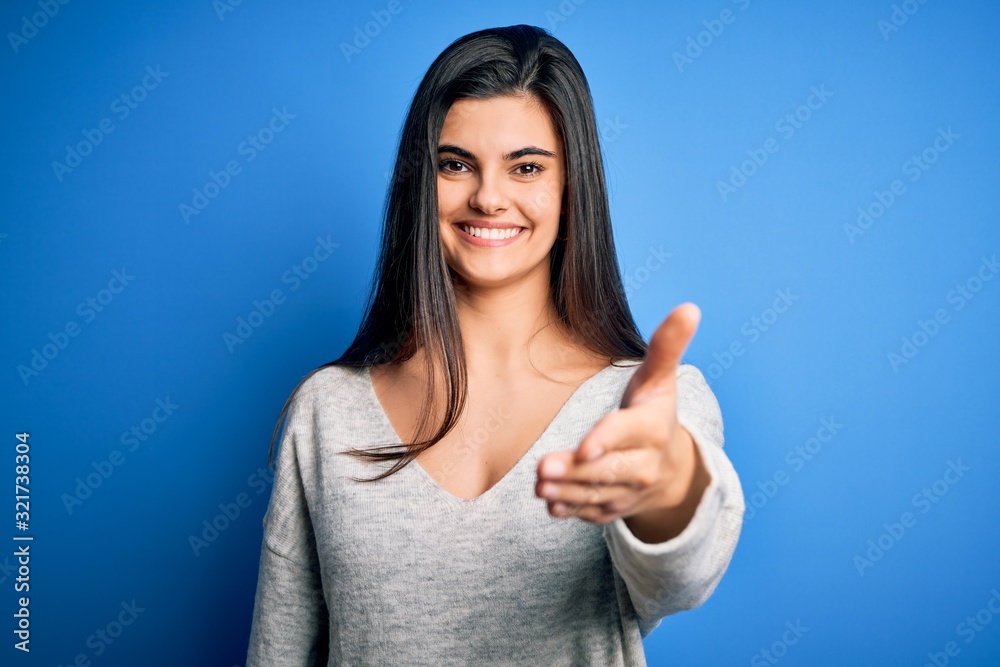 Young beautiful brunette woman wearing casual sweater standing over blue background smiling friendly offering handshake as greeting and welcoming. Successful business. Young beautiful brunette woman wearing casual sweater standing over blue background smiling friendly offering handshake as greeting and welcoming. Successful business.