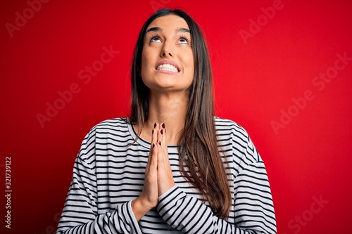 Young beautiful brunette woman wearing casual striped t-shirt over red background begging and praying with hands together with hope expression on face very emotional and worried. Begging.