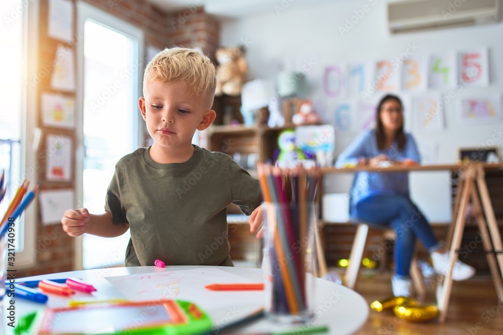 Fototapeta premium Young caucasian child playing at playschool with teacher. Mother and son at playroom drawing a draw with color pencils, young woman at the background sitting on desk.